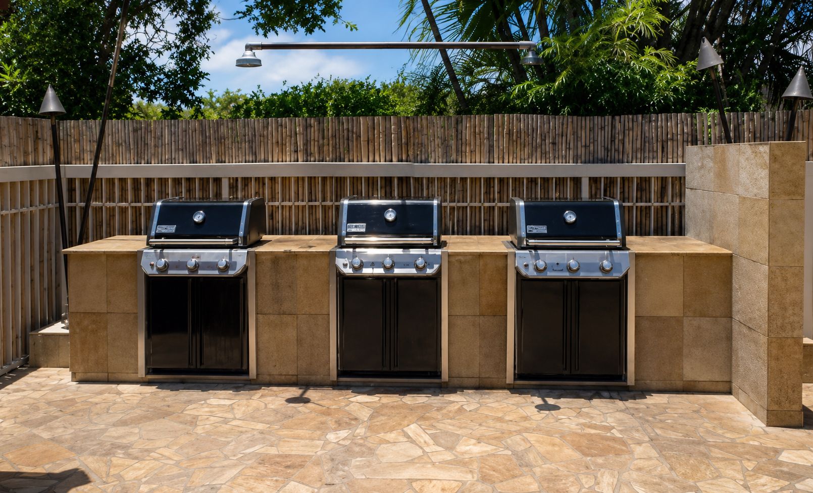 Three barbecue grills with a stone-tiled patio and a wooden fence in the background.