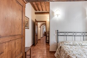A bedroom with a wooden wardrobe, a bed with floral bedding, a white wall with a light fixture, and a tiled floor.