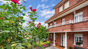 Une maison en briques rouges avec un balcon, un jardin où poussent des rosiers et un ciel d’un bleu limpide.