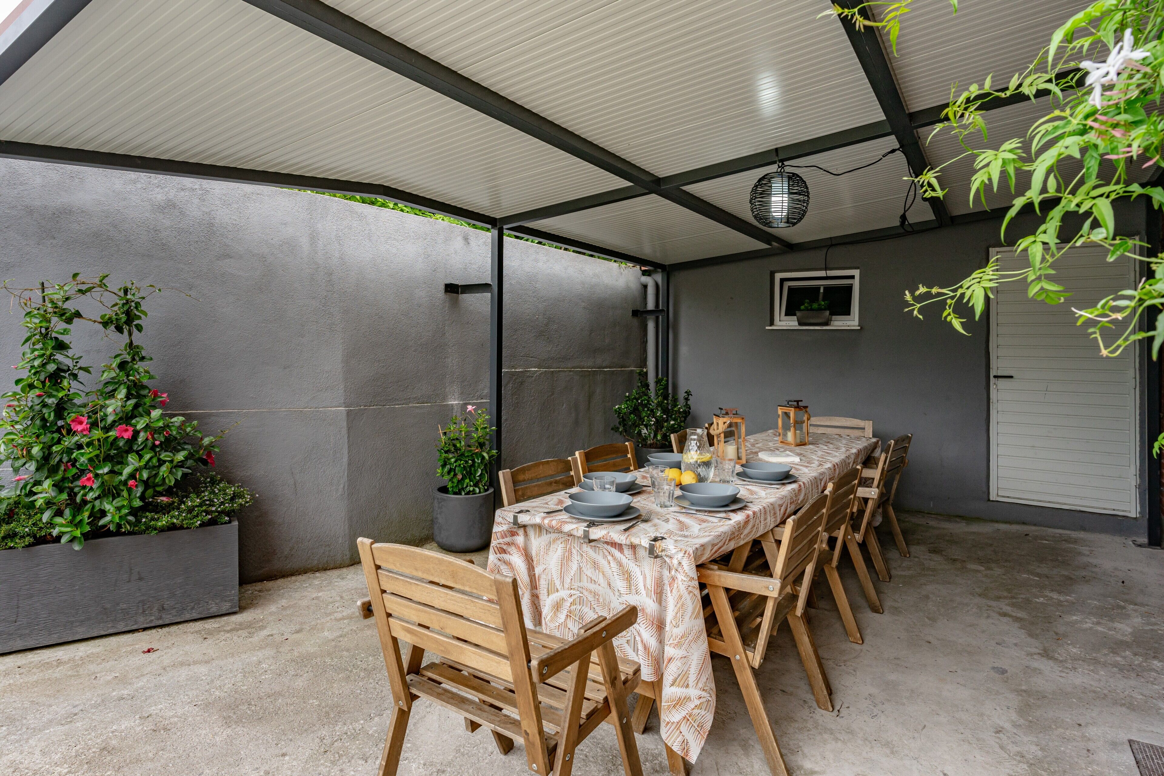 Un espacio cubierto para comer al aire libre con una mesa larga, sillas, plantas en macetas y una lámpara colgante.