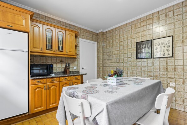 Une cuisine avec des meubles de rangement en bois, un réfrigérateur blanc, un four à micro-ondes et une table à manger recouverte d’une nappe.