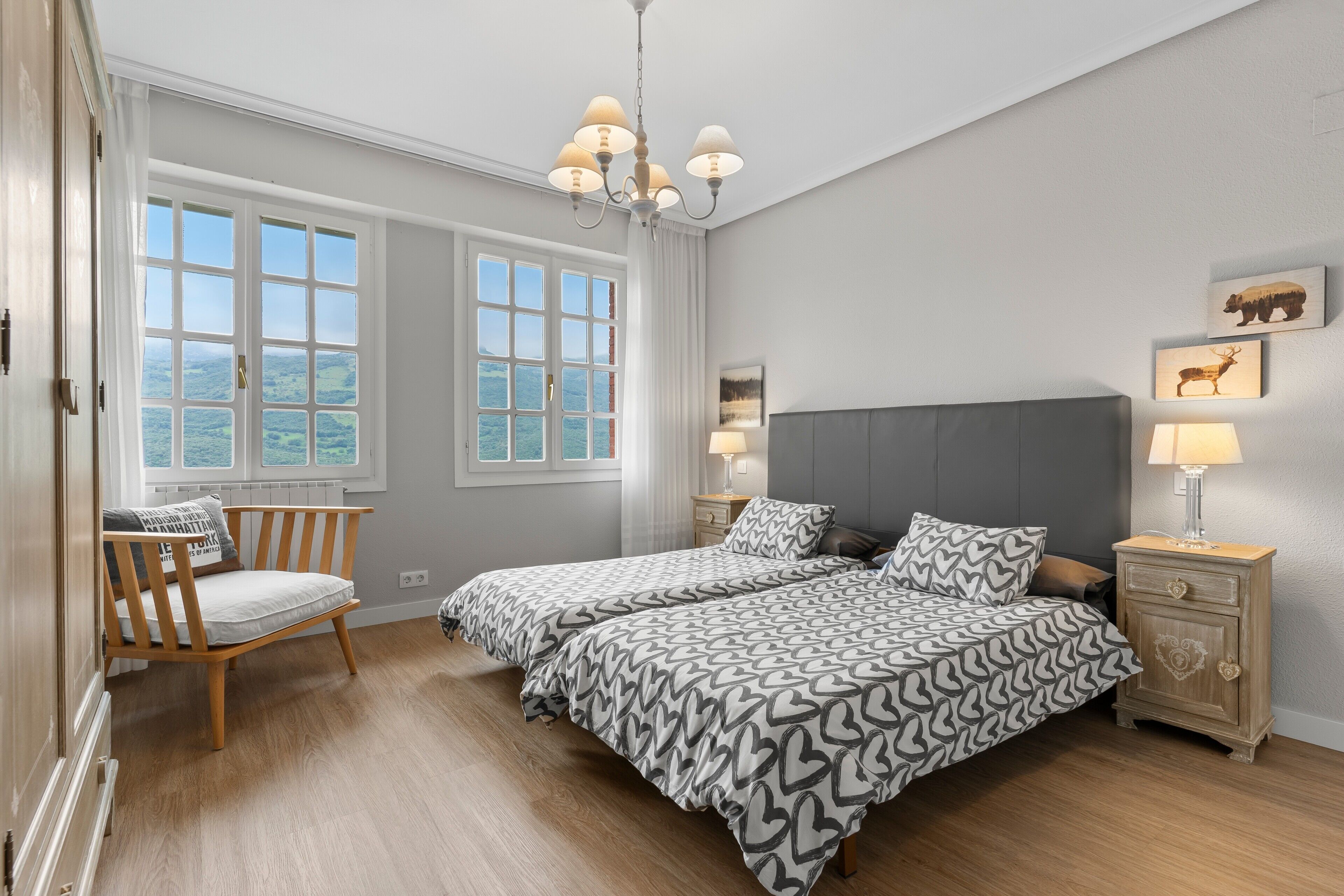 Un dormitorio con dos camas, una silla de madera, una mesita de noche con lámpara y vistas al exterior.