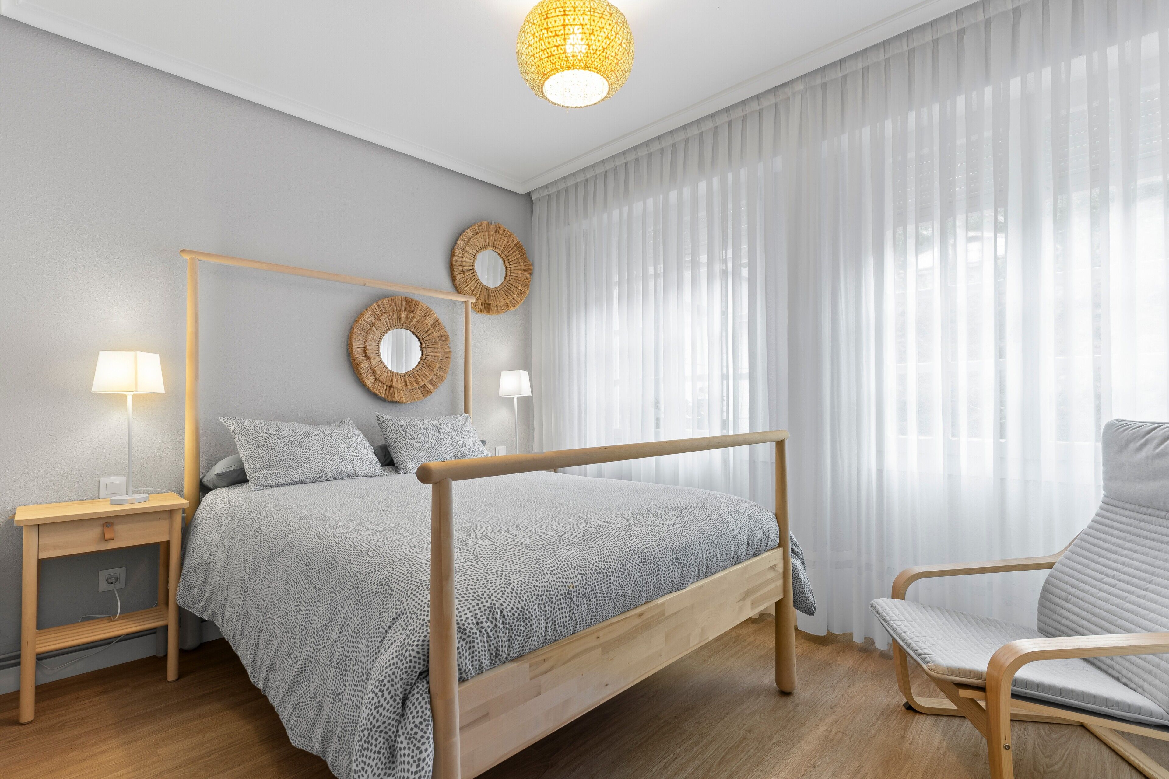 Un dormitorio moderno con cama de madera, lámparas de noche, una silla y un ventanal con cortinas translúcidas.