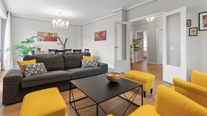 Un salon moderne avec un canapé d’angle, une table basse et des fauteuils jaunes. On aperçoit à l’arrière-plan un coin repas avec une table et des chaises.