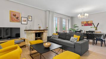 A living room with a sofa, yellow armchairs, a TV, a fireplace, and a dining area with a table and chairs.