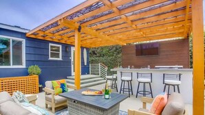 Un espacio de estar al aire libre cubierto con una pérgola, una barra con taburetes y una mesa de comedor con sillas.