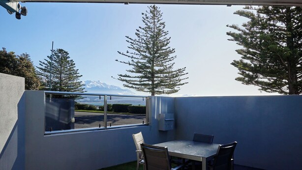 A covered patio with a table and chairs, a view of a mountain, and tall evergreen trees. - Kaikoura Luxury Apartments (Kaikoura)