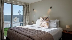 A hotel room with a bed, a nightstand, a lamp, and a view of a building through a window. - Kaikoura Luxury Apartments (Kaikoura)