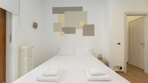 Ein schlicht eingerichtetes Hotelzimmer mit einem ordentlich bezogenen Bett, einem Heizkörper, einem kleinen Nachttisch mit einer Lampe und einer Tür zu einem weiteren Raum.