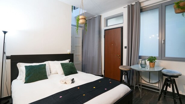 Habitación de hotel ordenada con una cama, una mesita redonda pequeña, una silla, una ventana con cortinas y una planta colgante.