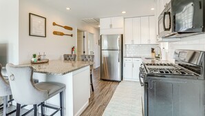 A modern kitchen with white cabinets, a stainless steel refrigerator, a gas stove, and a microwave. There are two bar stools and a framed picture on the wall. - Little Egg Harbor Township Retreat w/ Private Dock (Little Egg Harbor Township)