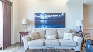 Una sala de estar con un sofá color crema, dos mesitas de noche con lámparas y un cuadro de gran tamaño con vista al océano.