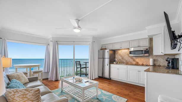 A modern living room with a view of the ocean, a kitchen with white cabinets and stainless steel appliances, and a ceiling fan. - 🌴BBC 214 - Gulf Front Condo - Pool & Beach are OPEN! (Belleair Beach)