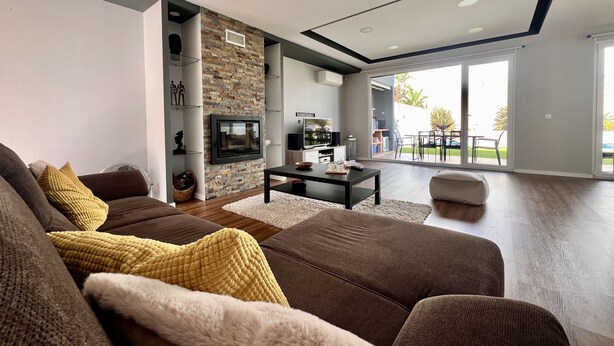 A modern living room with a stone accent wall, a flat-screen TV, and a dining area with a view to the outdoors. - Casa Giumeni - House with swimming pool (Fernão Ferro)
