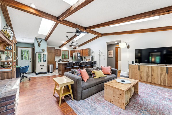 A spacious living area with a sofa, coffee table, and a TV. There is a kitchen in the background with wooden cabinets and a dining area.