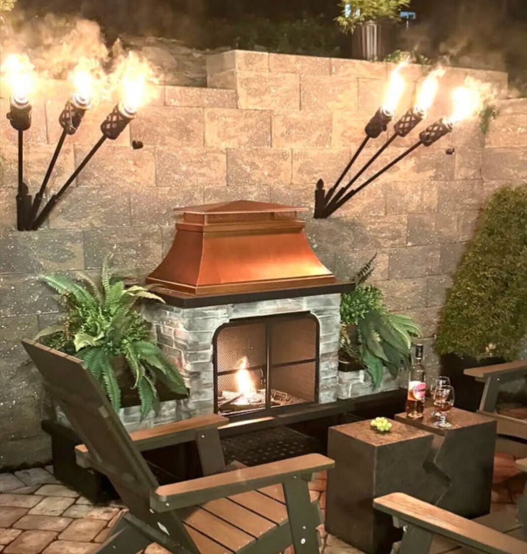 A stone fireplace with a metal hood, surrounded by chairs and potted plants.