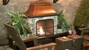 Una chimenea de piedra con capucha metálica, rodeada de sillones y plantas en macetas.