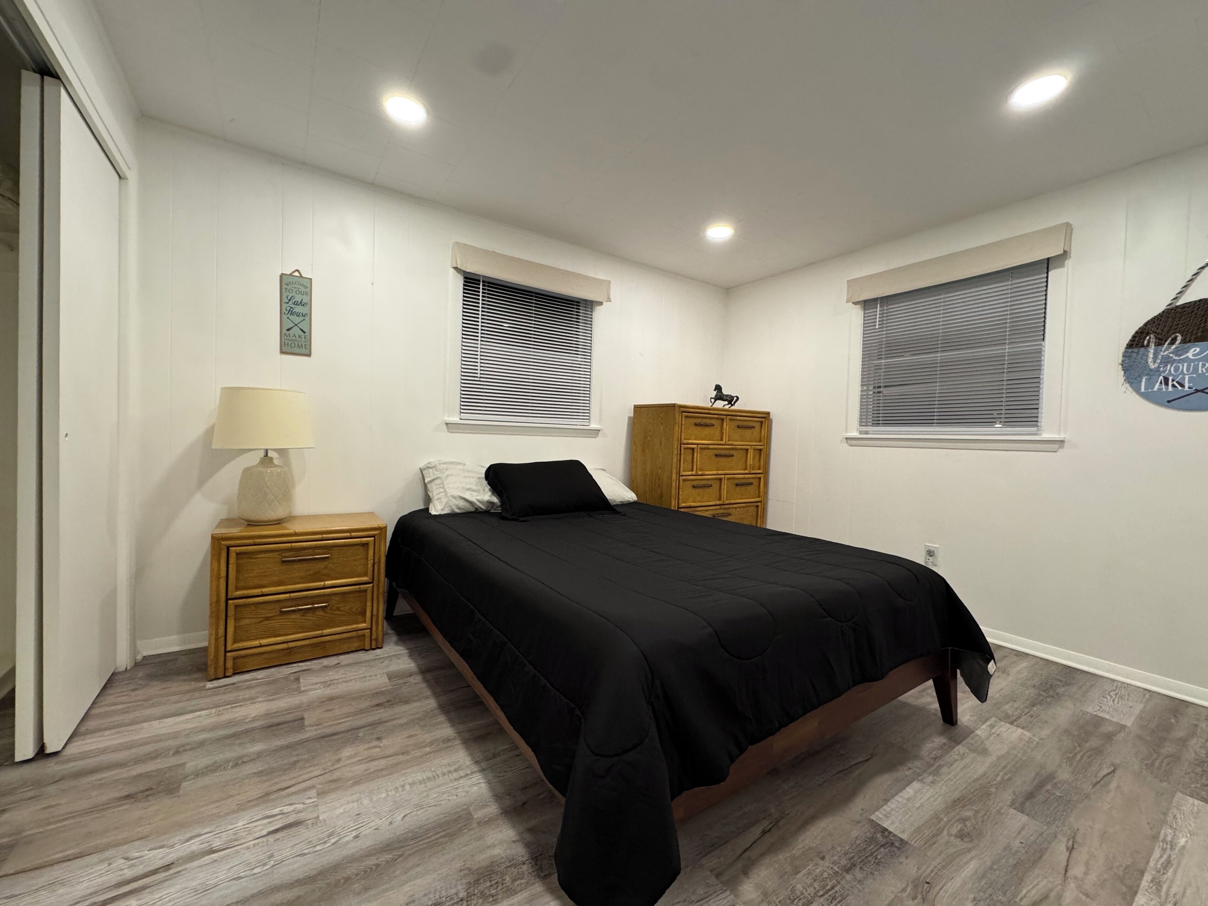 A bedroom with a bed, wooden nightstand, dresser, and two windows with blinds.