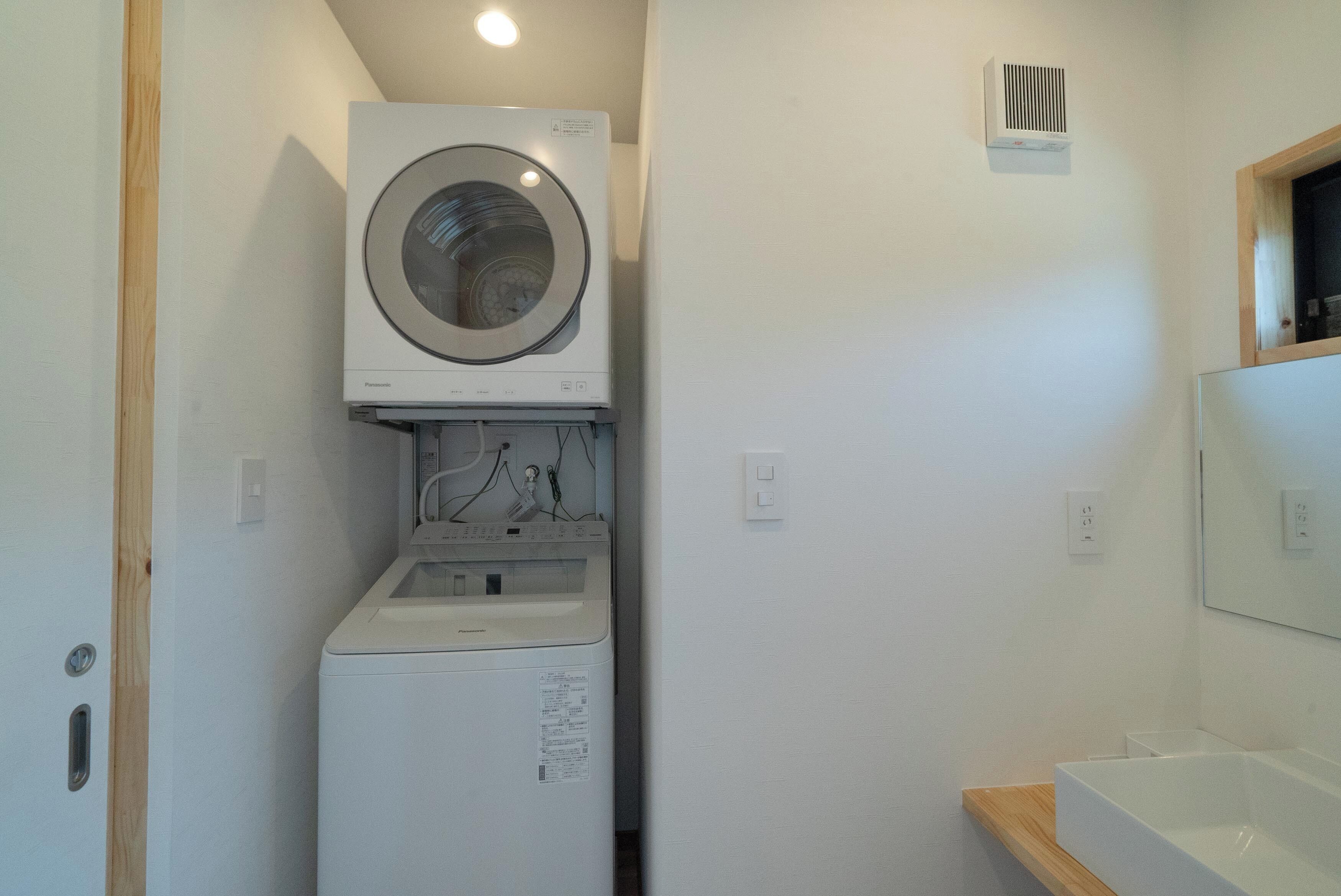 A compact laundry area with a front-loading washer and dryer, a wooden shelf, and a mirror above the sink.