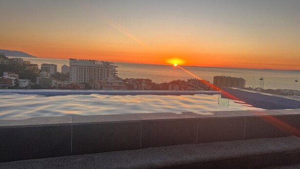 A sunset view from a balcony overlooking a coastal cityscape with buildings and a body of water.