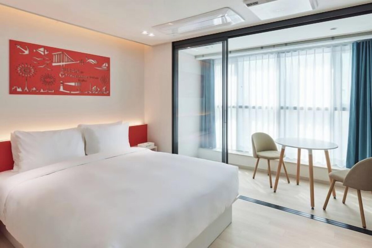 A modern hotel room with a large bed, a small table with chairs, and a large window with curtains.