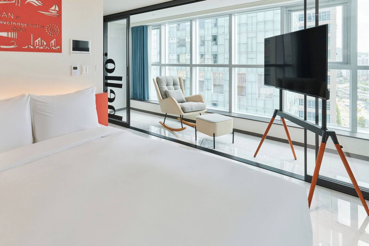 Moderni hotellihuone, jossa on suuri sänky, televisio, keinutuoli ja suuret ikkunat, joista avautuu näkymä kaupunkiin.