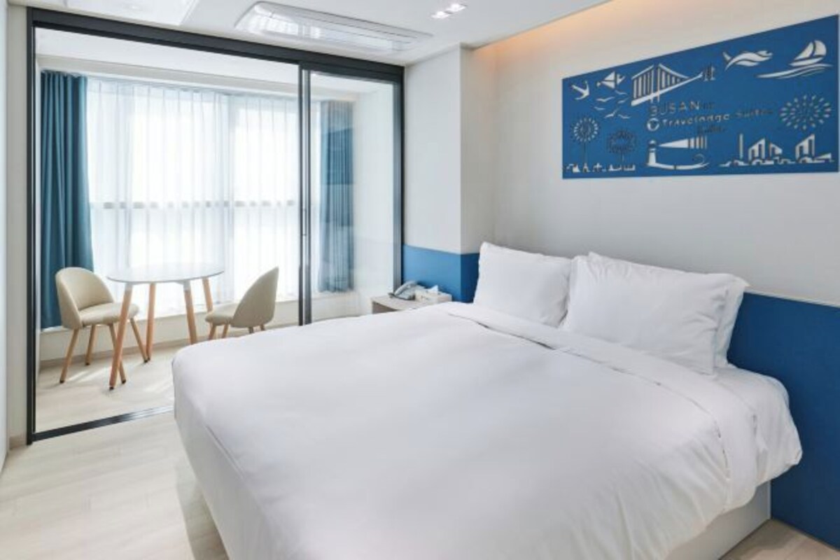 A hotel room with a large bed, a desk with chairs, and a view through a glass door.