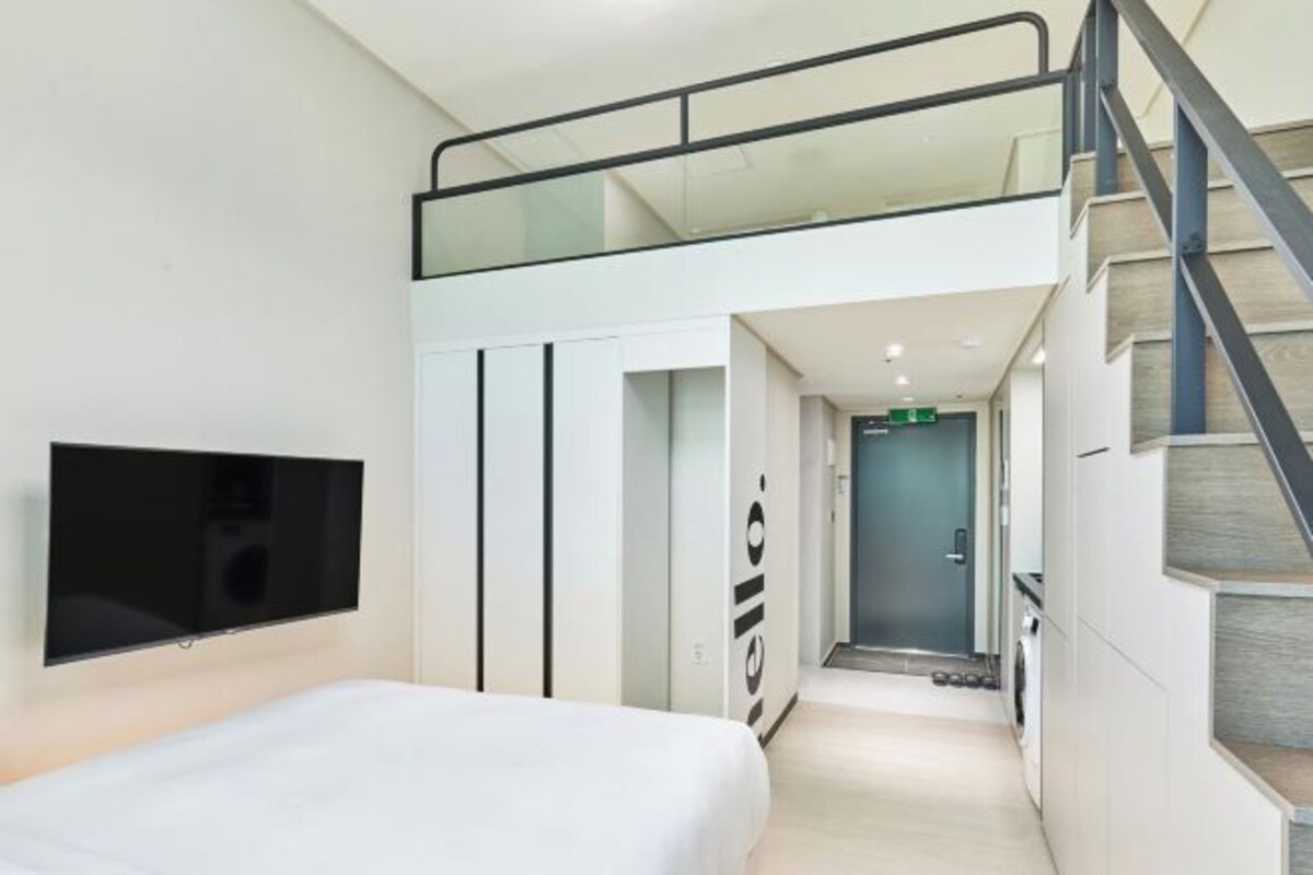 A modern hotel room with a bed, a television mounted on the wall, a staircase with a glass railing, and a door leading to a hallway.
