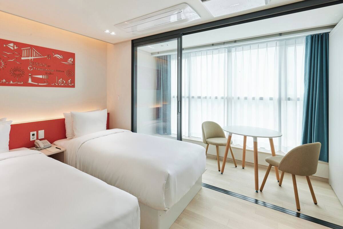 Hotellihuone, jossa on kaksi sänkyä, pieni pöytä tuoleineen, suuri ikkuna verhoilla ja punainen taideteos seinällä.