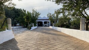 Un chemin pavé mène à un bâtiment doté d’un toit ondulé, entouré d’arbres et de murs blancs.