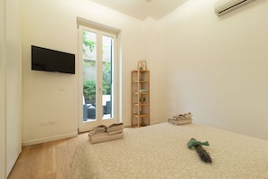 A bedroom with a bed, a TV mounted on the wall, a wooden shelf, and a window with a view of greenery.