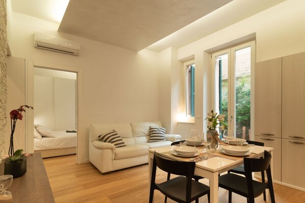 Una habitación moderna y bien iluminada con una mesa de comedor preparada para dos personas, un sofá y una cama al fondo.