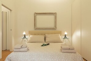 A bedroom with a bed, two bedside lamps, a framed picture on the wall, and folded towels on the bed.
