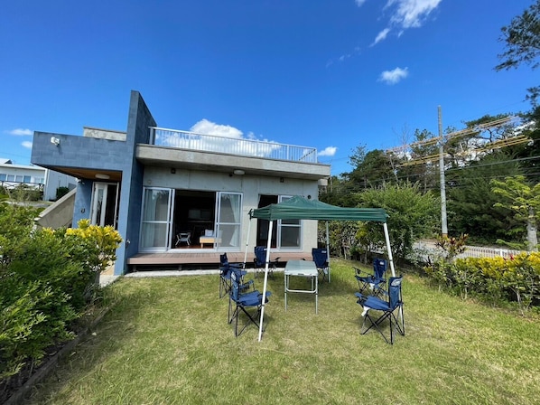A modern house with a balcony, a covered patio with chairs, and a grassy yard. - Tida Gesashi (Higashi)