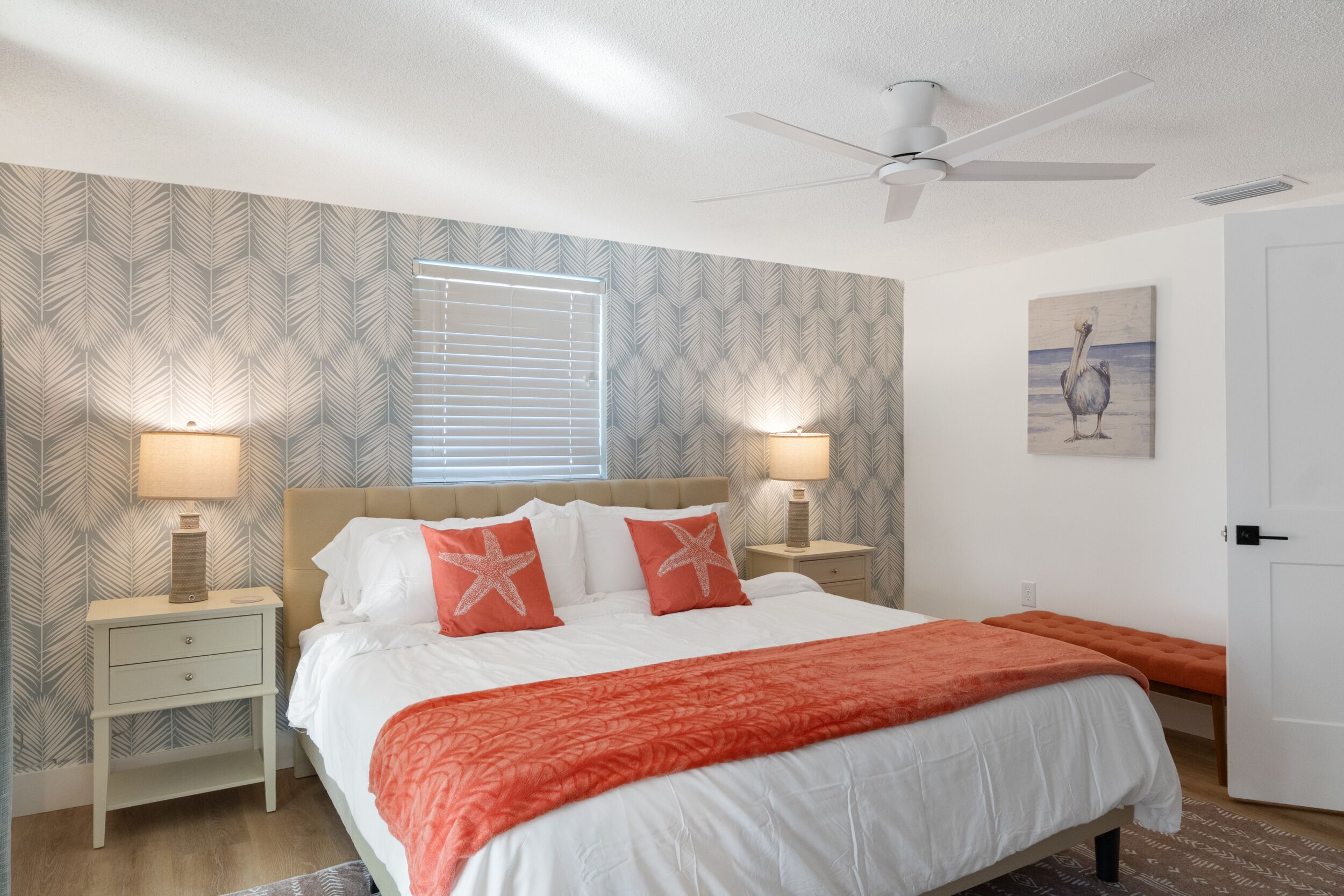 A bedroom with a bed, two bedside tables, a ceiling fan, a window with blinds, and a wall with a leaf pattern.