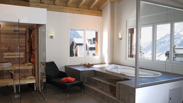 A modern bathroom with a wooden ceiling, a large mirror, a bathtub, and a window with a mountain view. - Apartment B01 by Interhome (Leukerbad)