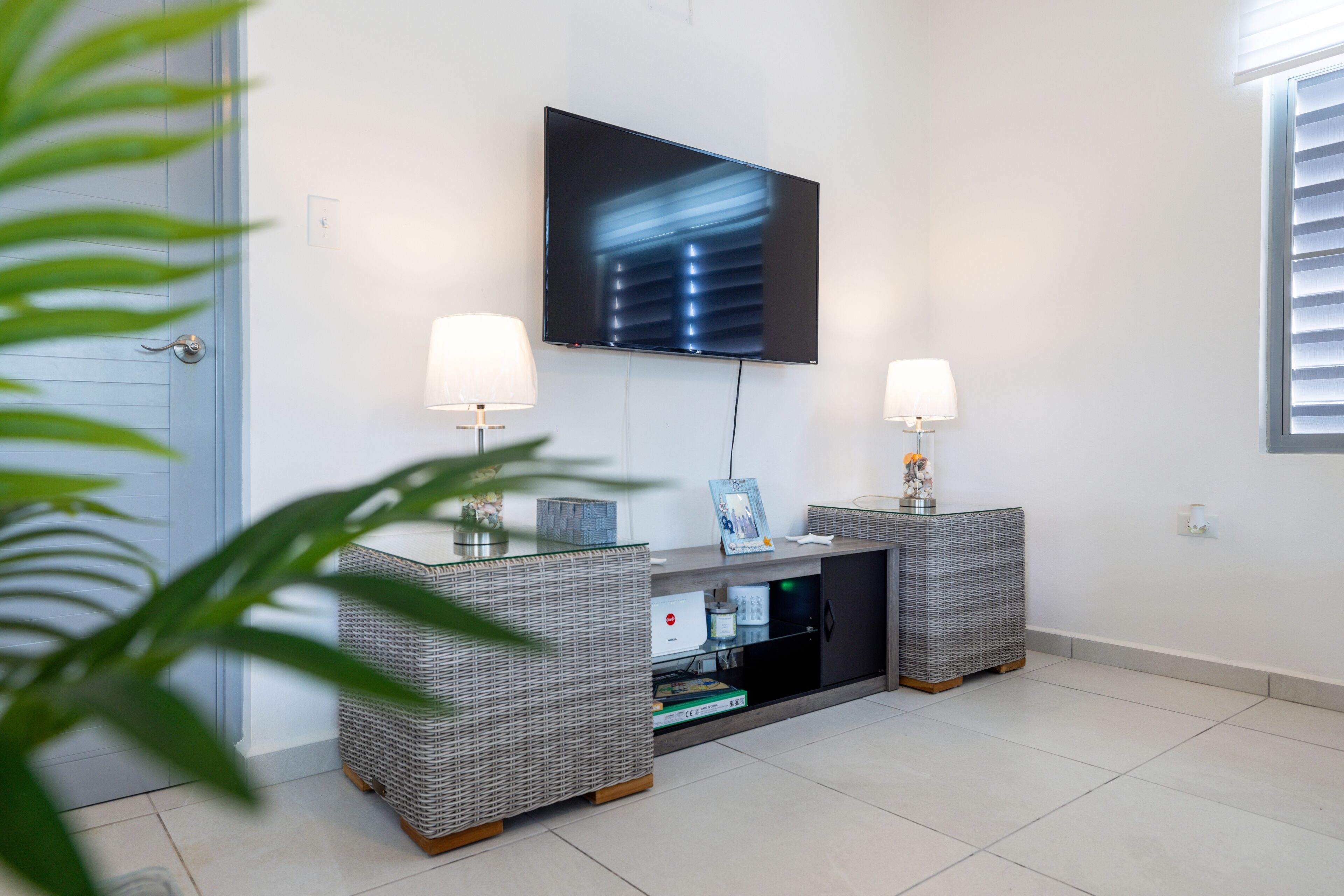 A modern living room with a flat-screen TV mounted on the wall, two side tables with lamps, and a wicker console table with a glass top.