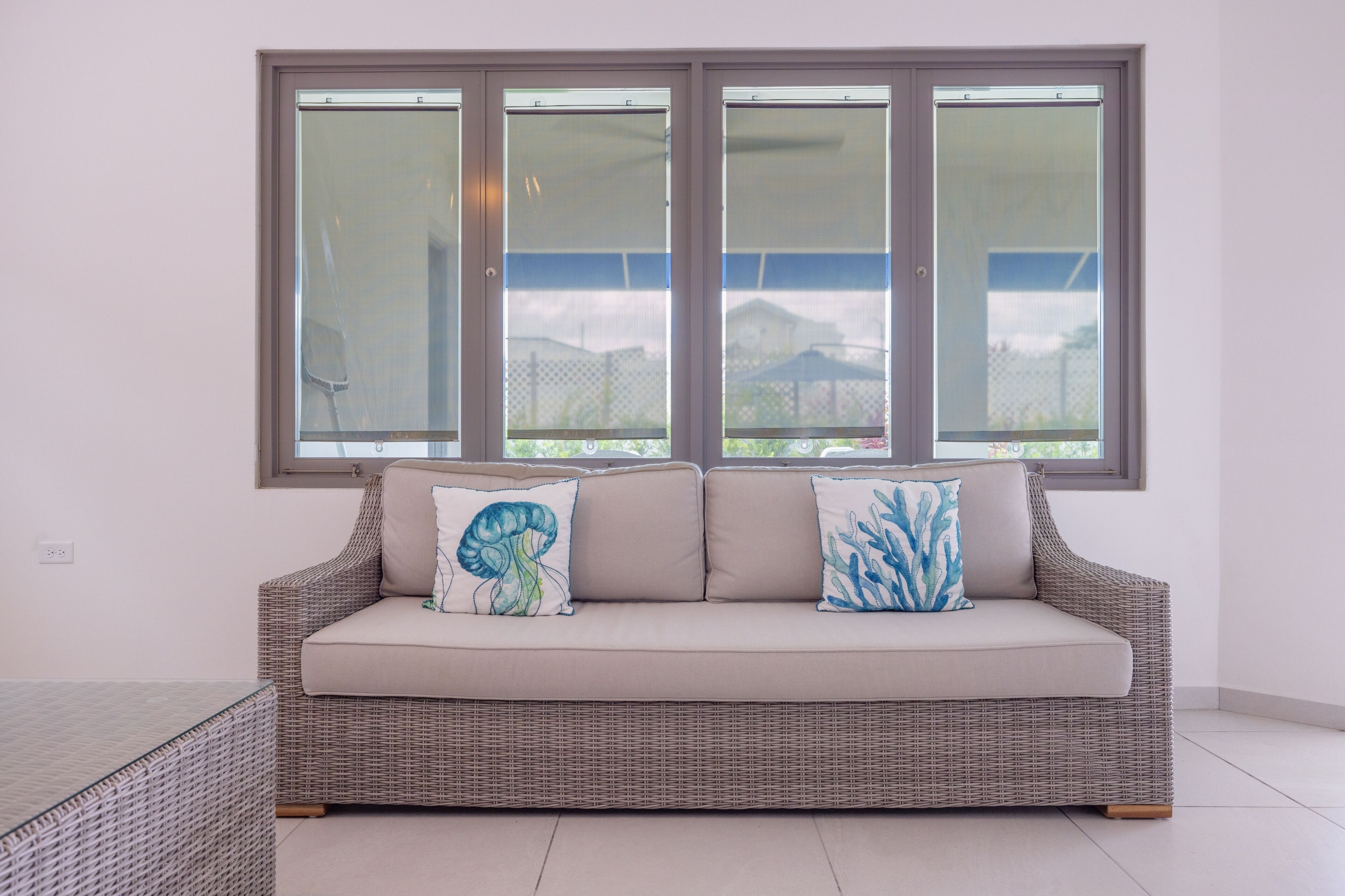 A wicker sofa with two decorative pillows, positioned in front of a large window with a view of a fence and mountains.