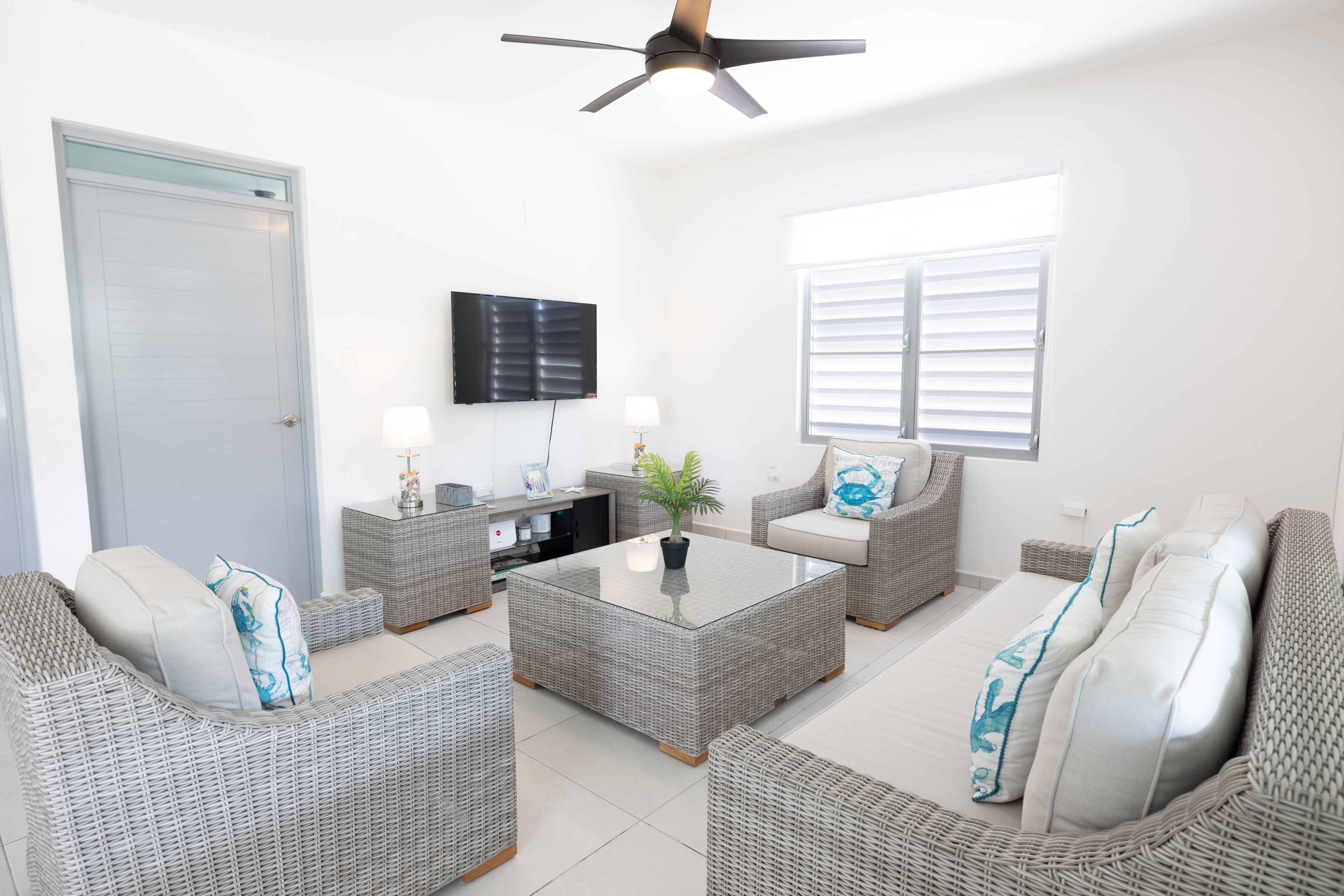 A modern living room with wicker furniture, a glass coffee table, and a ceiling fan.