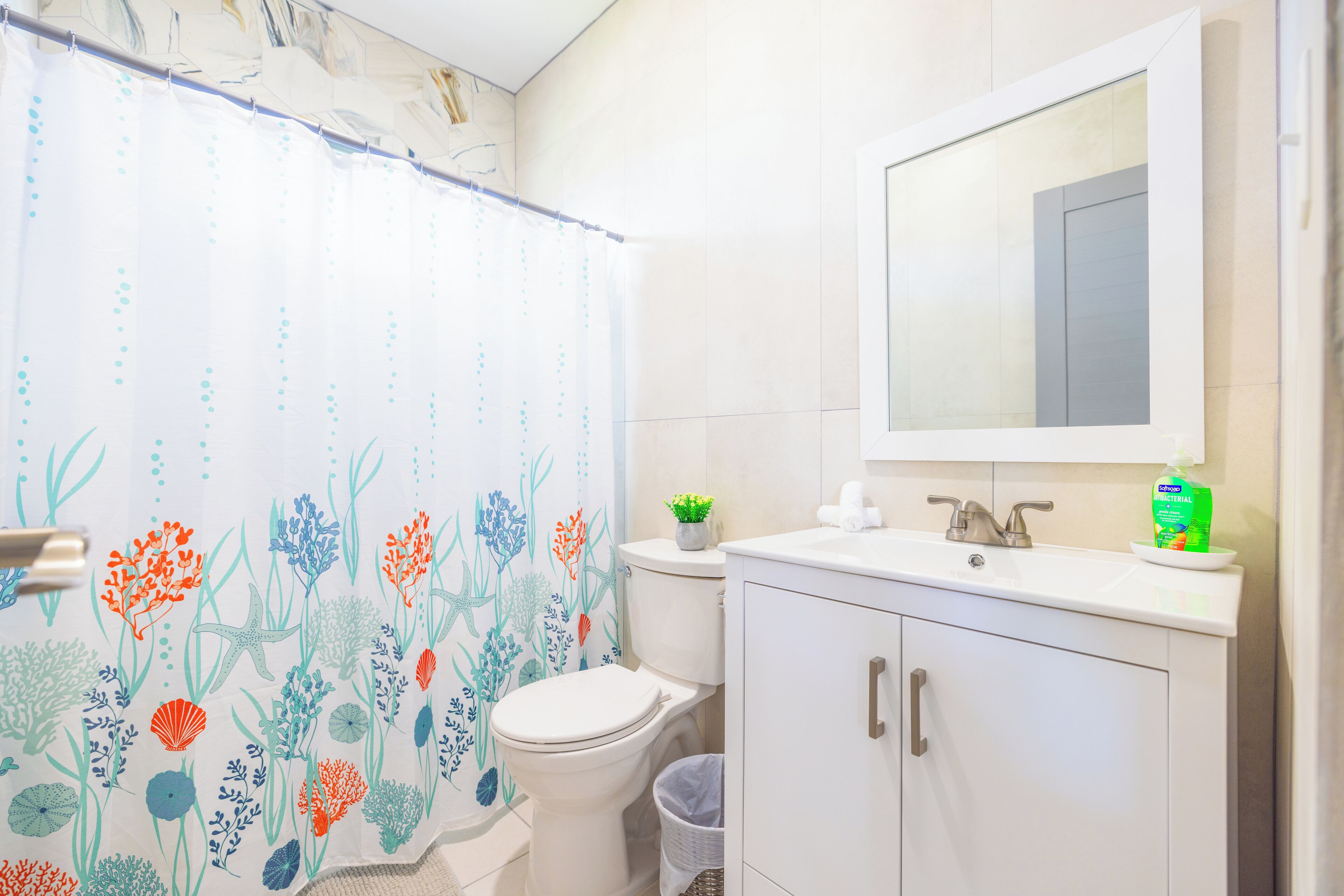 Une salle de bain avec un rideau de douche orné d’un motif floral, des toilettes, un lavabo avec un miroir au-dessus et une poubelle.