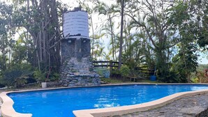 Une piscine bordée de pierres, avec une structure en pierre abritant un réservoir d’eau.