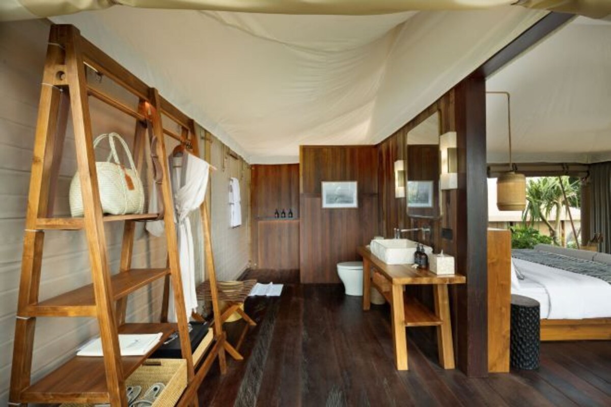 A room with a wooden floor, a bed with a white headboard, a wooden shelf with bags, a small table with toiletries, and a large mirror.