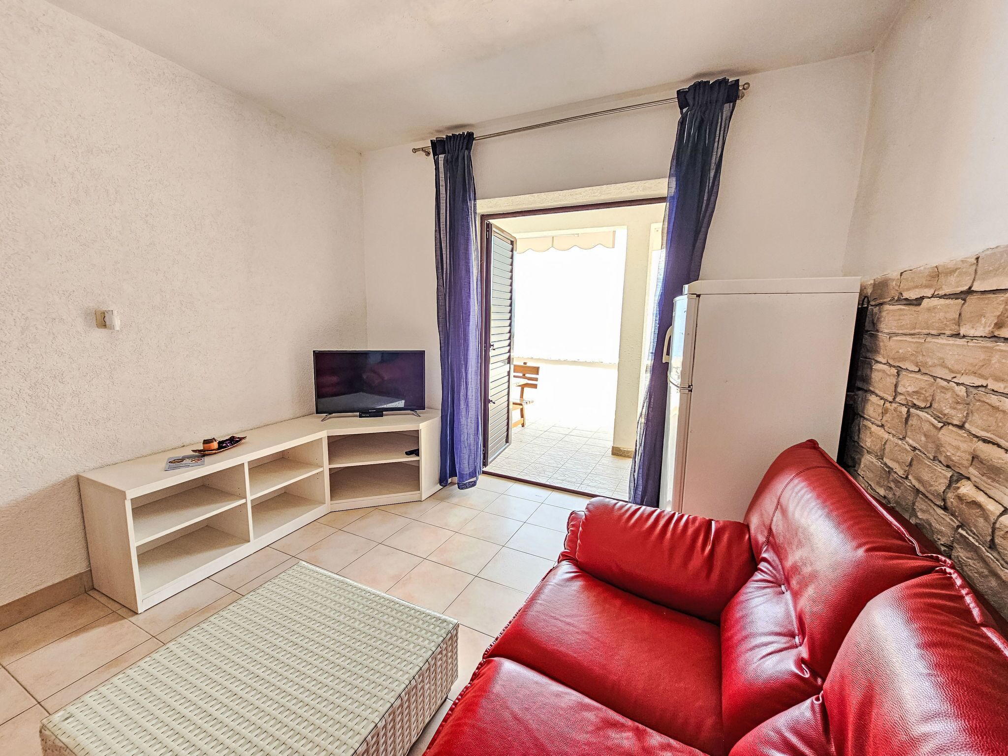 Un salon avec un canapé en cuir rouge, un téléviseur posé sur un meuble blanc, un pouf en osier et un mur en pierre.