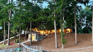 Une propriété au bord du lac, comprenant un chalet en bois, un mur de soutènement en pierre et un pont bleu qui enjambe l’eau.