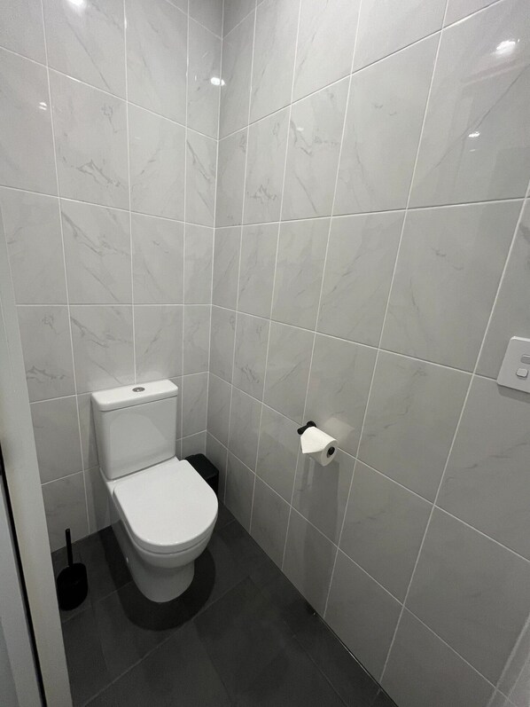 Une petite salle de bain moderne, équipée de toilettes, d’un drain noir au sol et d’un porte-rouleau de papier toilette fixé au mur.