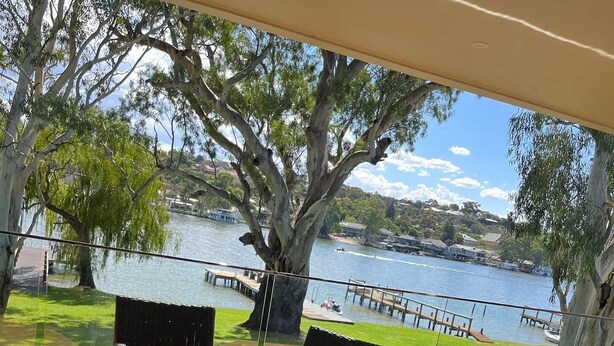 An outdoor dining area with a wooden table and wicker chairs, overlooking a lake with a dock and boats.