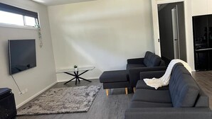 A modern living room with a sectional sofa, a flat-screen TV mounted on the wall, a small table with a glass top, and a dark wood floor.