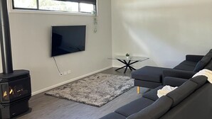 A modern living room with a large grey sofa, a flat-screen TV mounted on the wall, a glass coffee table, and a black wood-burning stove.