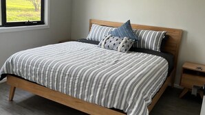 A bedroom with a wooden bed, a nightstand, a window with a view of greenery, and a ceiling vent.