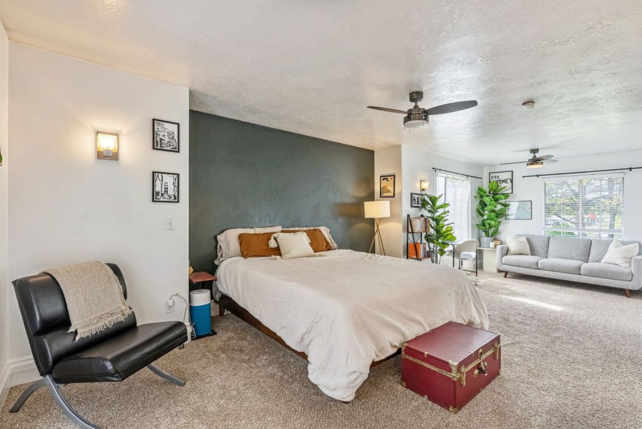 A bedroom with a bed, a chair, a suitcase, a ceiling fan, and a window.